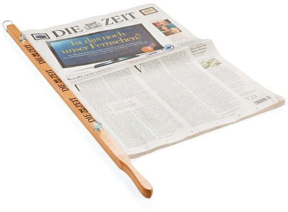 Ein Zeitungshalter aus Holz, der eine aufgeklappte Ausgabe der Zeitung 'Die Zeit' hält. Das Gerät besteht aus einem langen Holzstab mit Metallklammern, die die Zeitung an Ort und Stelle halten.