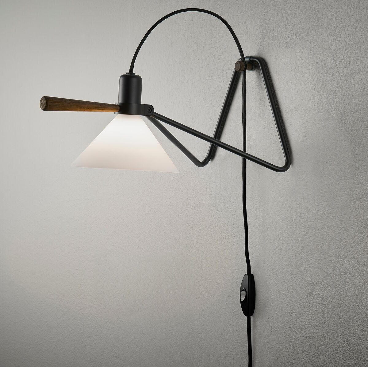 Eine moderne Tisch- oder Hängelampe mit einem schwarzen Metallgestell und einem weißen, konischen Lampenschirm, montiert an der Wand. Die Lampe hat ein minimalistisches Design mit einem Hängeschalter an einem schwarzen Kabel.