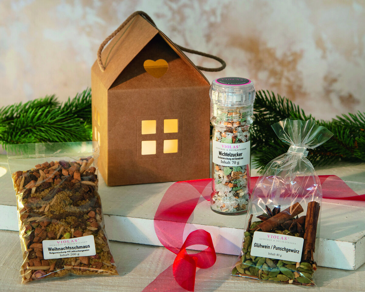 Ein Süsses Weihnachtshaus Geschenkset bestehend aus einem beleuchteten Papphaus, Weihnachtsdecken, einer Gewürzmischung und Weihnachtstee-Verpackungen, verziert mit einer roten Schleife, und im Hintergrund Tannenzweige.