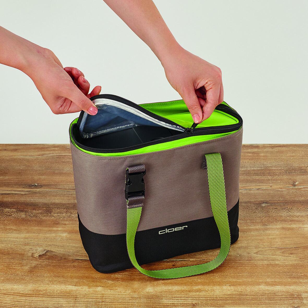 Person öffnet eine grau-grüne Lunch-Tasche mit Reißverschluss auf einem Holztisch.