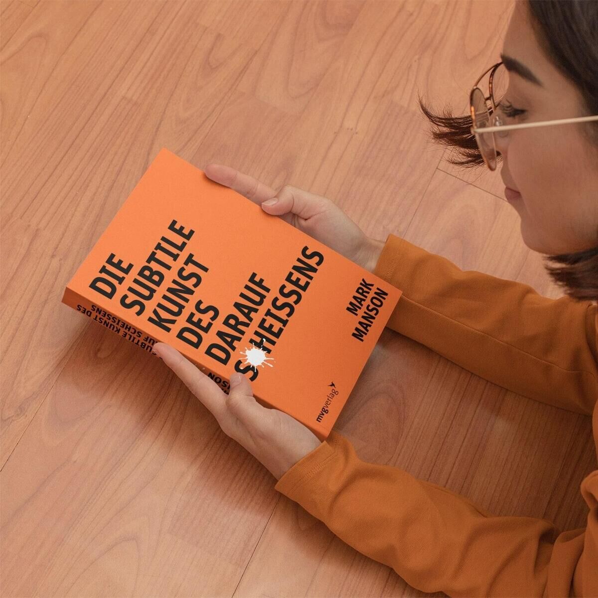 Eine Frau mit Brille liegt auf einem Holzboden und hält das Buch 'Die subtile Kunst des darauf Scheißens' von Mark Manson in den Händen. Das Buch hat ein orangefarbenes Cover mit schwarzem Text.