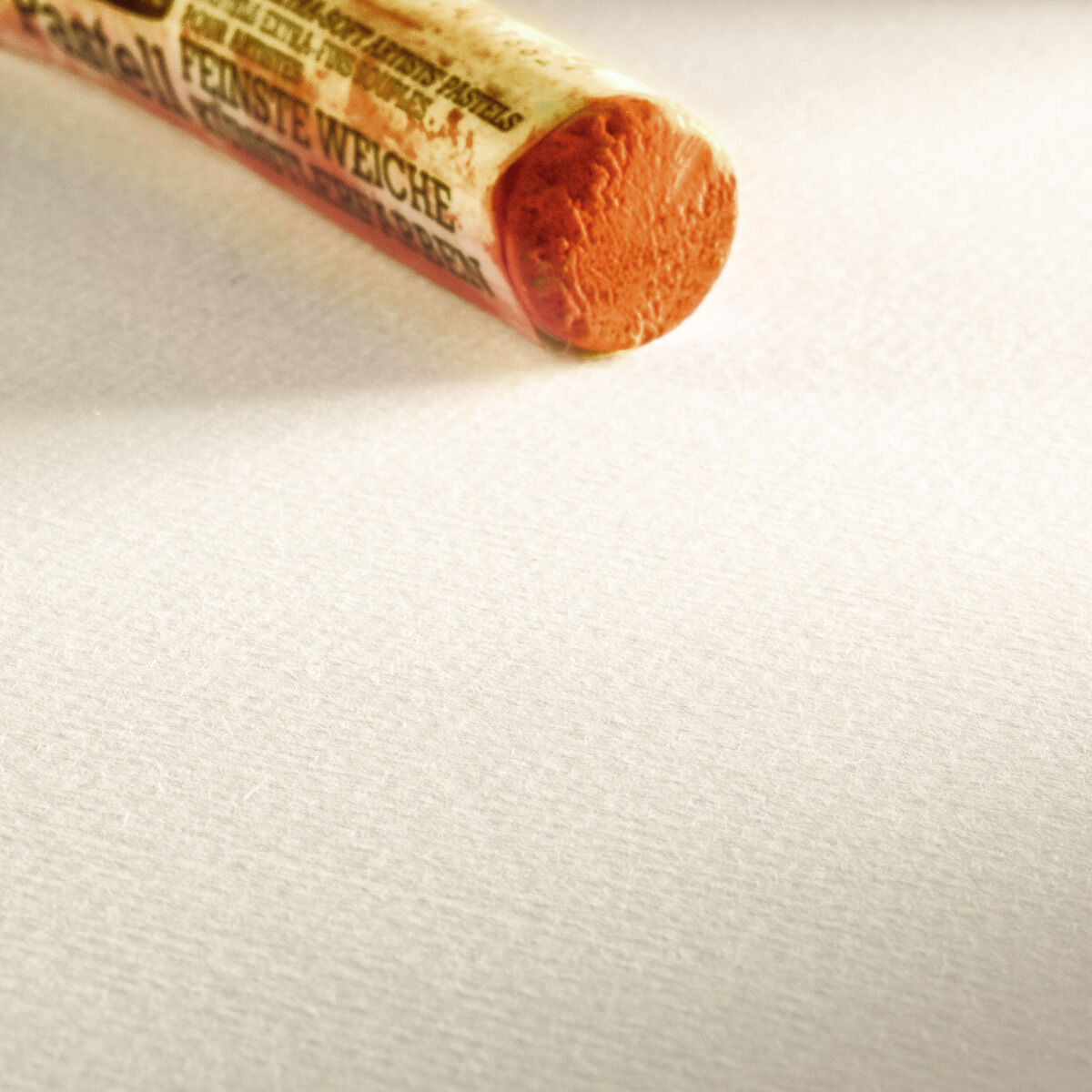 Ein weißes Blatt Skizzenpapier mit einer orangefarbenen Pastellkreide steht auf der linken Seite des Bildes. Die Oberfläche des Papiers ist leicht texturiert.