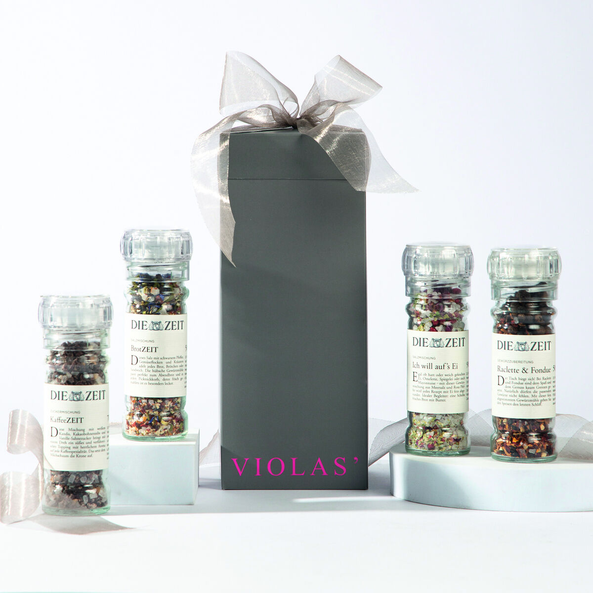 Das Bild zeigt ein 4er Gewürzmühlen-Set von ZEIT in einer eleganten Geschenkverpackung. Die verpackung ist schwarz mit einer edlen Schleife verziert und die Gewürzmühlen enthalten unterschiedliche farbenfrohe Gewürzmischungen. Auf der Verpackung ist der violette Schriftzug 'Violas*' zu sehen.