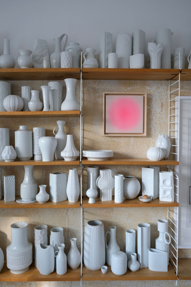 Ein Regal mit weißen Vasen unterschiedlicher Formen und Größen. An der Wand hängt ein quadratisches Kunstwerk in einem Holzrahmen, das einen pinken Farbverlauf zeigt.