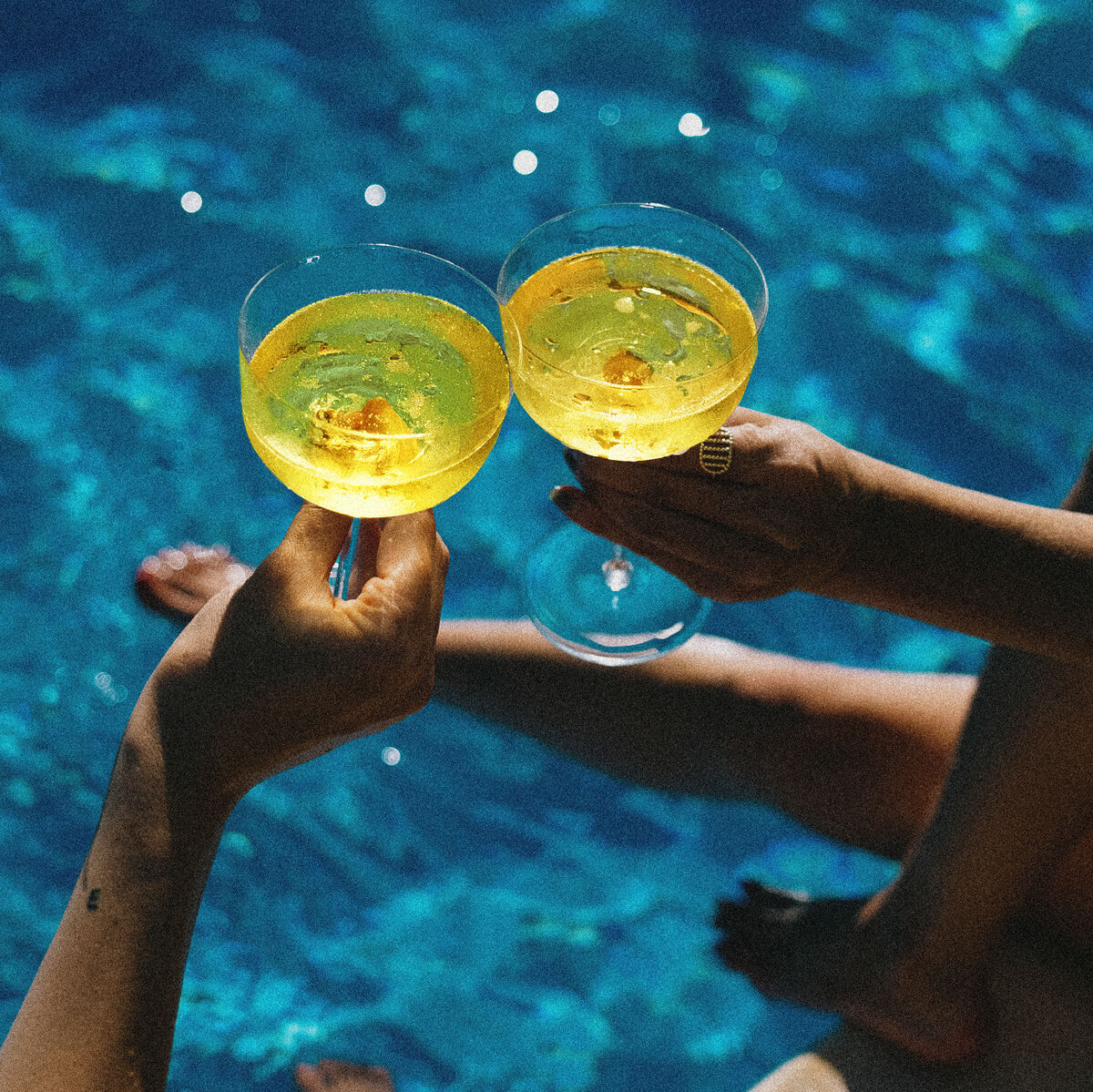 Zwei Personen stoßen mit Aperitif-Gläsern an, die ein gelbes Getränk enthalten, während sie am Rand eines klaren, türkisfarbenen Pools sitzen. Im Hintergrund sind der Pool und das Wasser zu sehen.