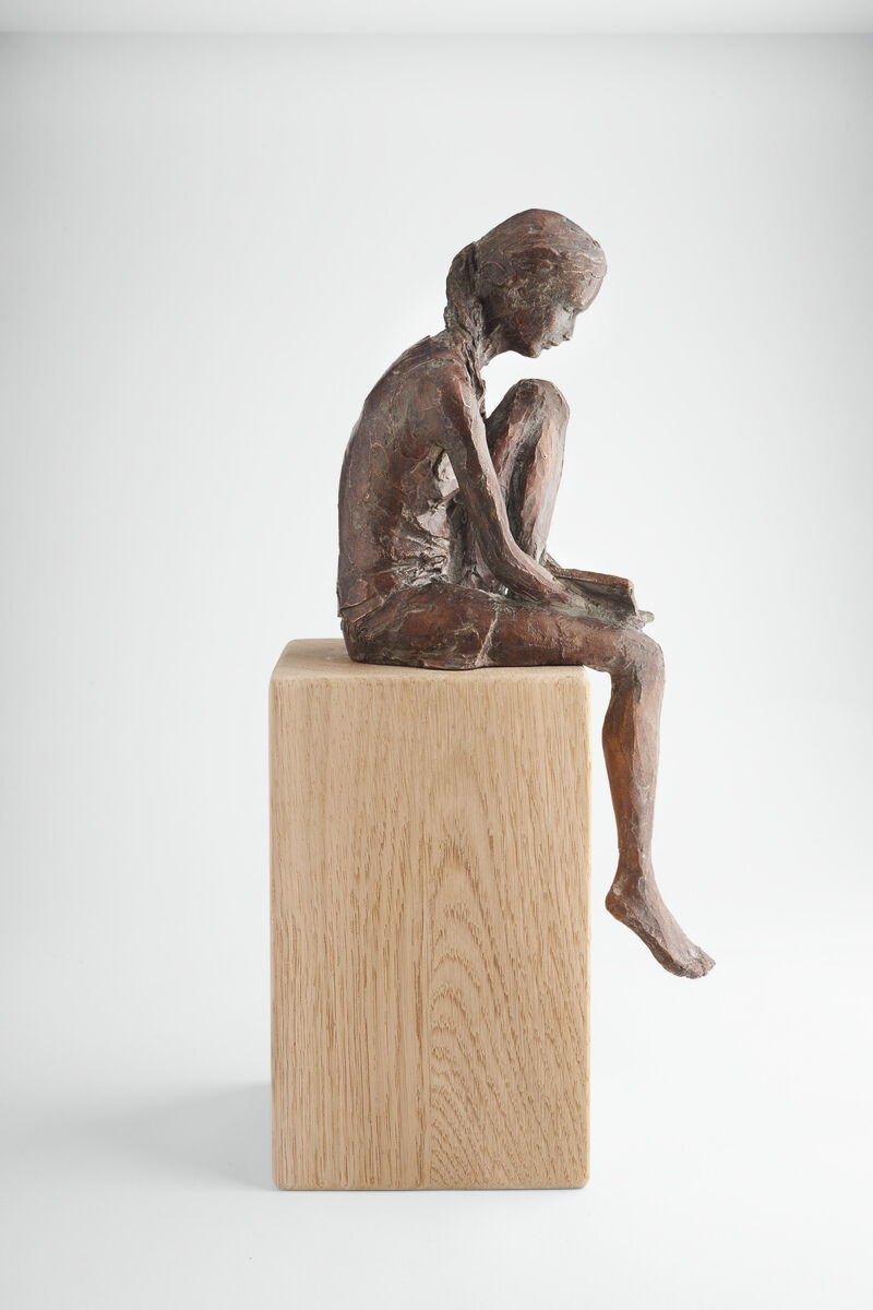 Die Skulptur 'Lesende' von Valerie Otte zeigt eine abstrakte, bronzene Figur eines sitzenden Mädchens, das in ein Buch vertieft ist. Die Figur sitzt auf einem rechteckigen Holzsockel aus heller Eiche. Der Hintergrund ist weiß und schlicht, wodurch die Skulptur hervorgehoben wird.