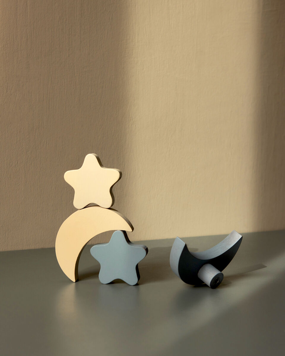 Eine minimalistische Fotografie zeigt drei an der Wand lehnende Haken in Form von zwei Sternen und einem Mond, die aus Holz gefertigt und in pastellfarbenen Tönen (hellblau, beige und dunkelgrau) bemalt sind.