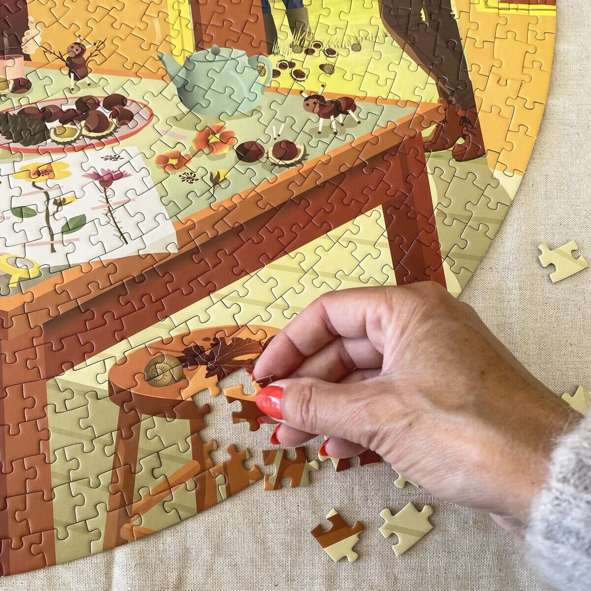 Nahaufnahme eines runden 1000-Teile-Puzzles »Chestnut Season« von VISSEVASSE. Eine Hand setzt ein Puzzlestück an, Motiv zeigt herbstliche Szene mit Tisch, Gebäck, Blumen, und Menschen in warmer Kleidung, sanfte warme Farbtöne dominieren das Bild.