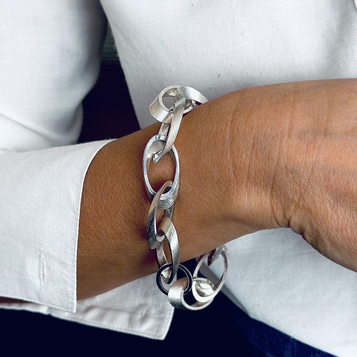 Ein silbernes Armband mit ovalen Gliedern, getragen auf dem Handgelenk einer Person, die ein weißes Hemd trägt. Das Armband hat ein elegantes und zeitloses Design.