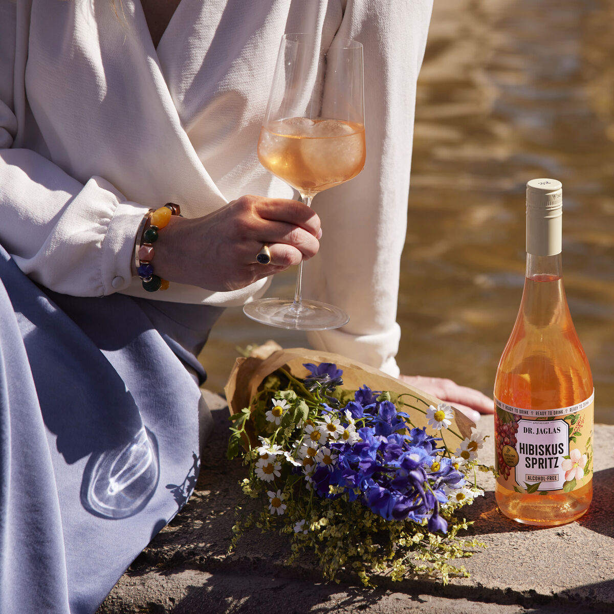 Eine Flasche Hibiskus Spritz steht neben Blumen und einem Glas Wein im Freien. Zwei Personen sind im entspannten Gespräch, die Szene wirkt sommerlich und gemütlich. Eine Schale mit Trauben ist ebenfalls sichtbar.