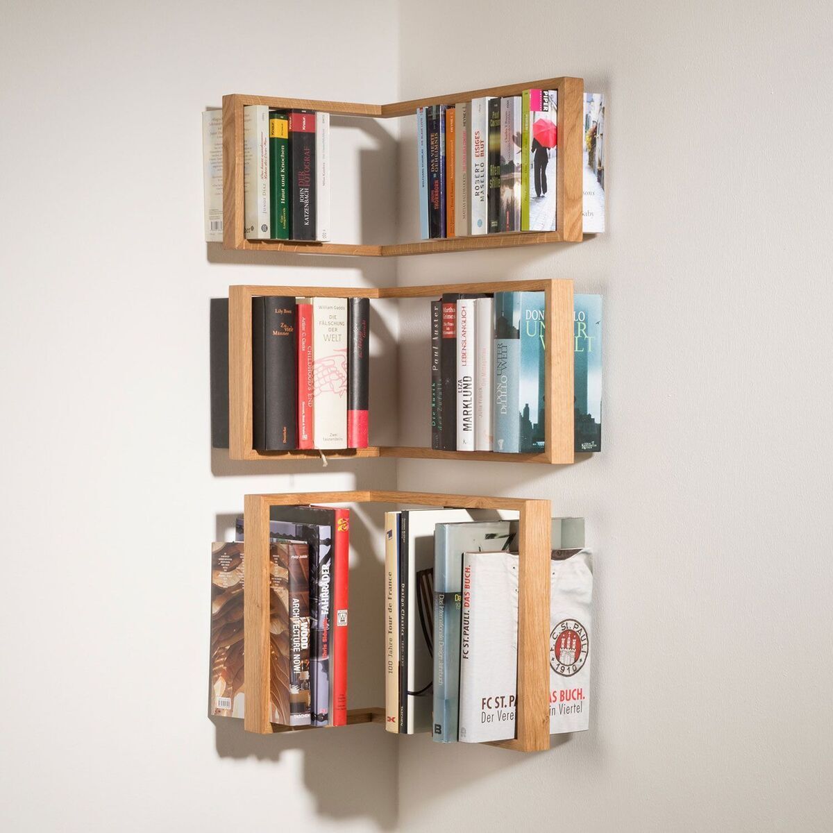 Ein Wandregal in Holzoptik, das in die Ecke montiert ist und mit verschiedenen Büchern gefüllt ist. Es besteht aus drei Ebenen, die jeweils eine unterschiedliche Anordnung von Büchern aufweisen.