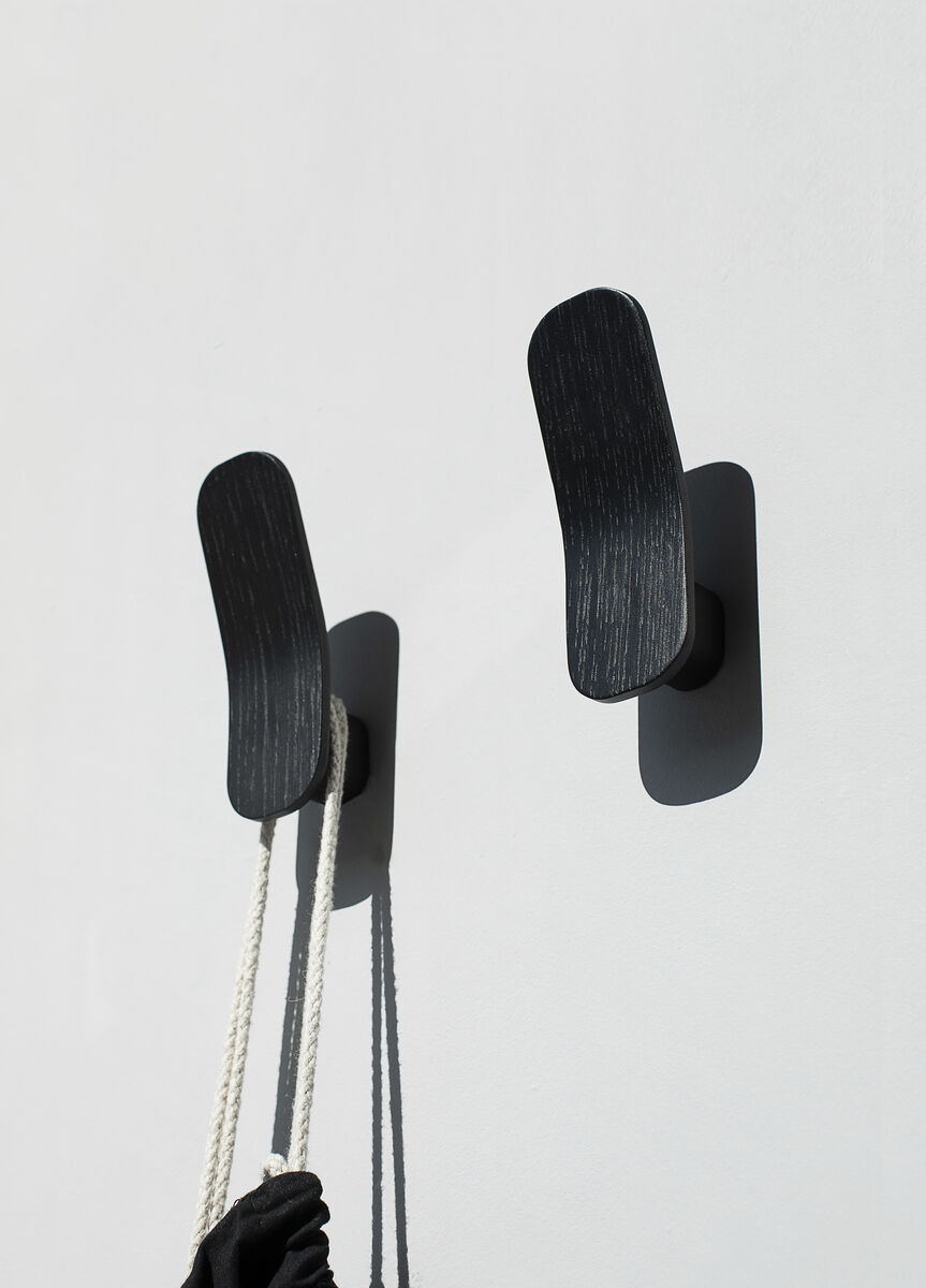 Zwei moderne Kleiderhaken »Eshaku« aus schwarzer Eiche, die an einer weißen Wand montiert sind, mit einem Seil und einem schwarzen Gegenstand, der an einem der Haken hängt.