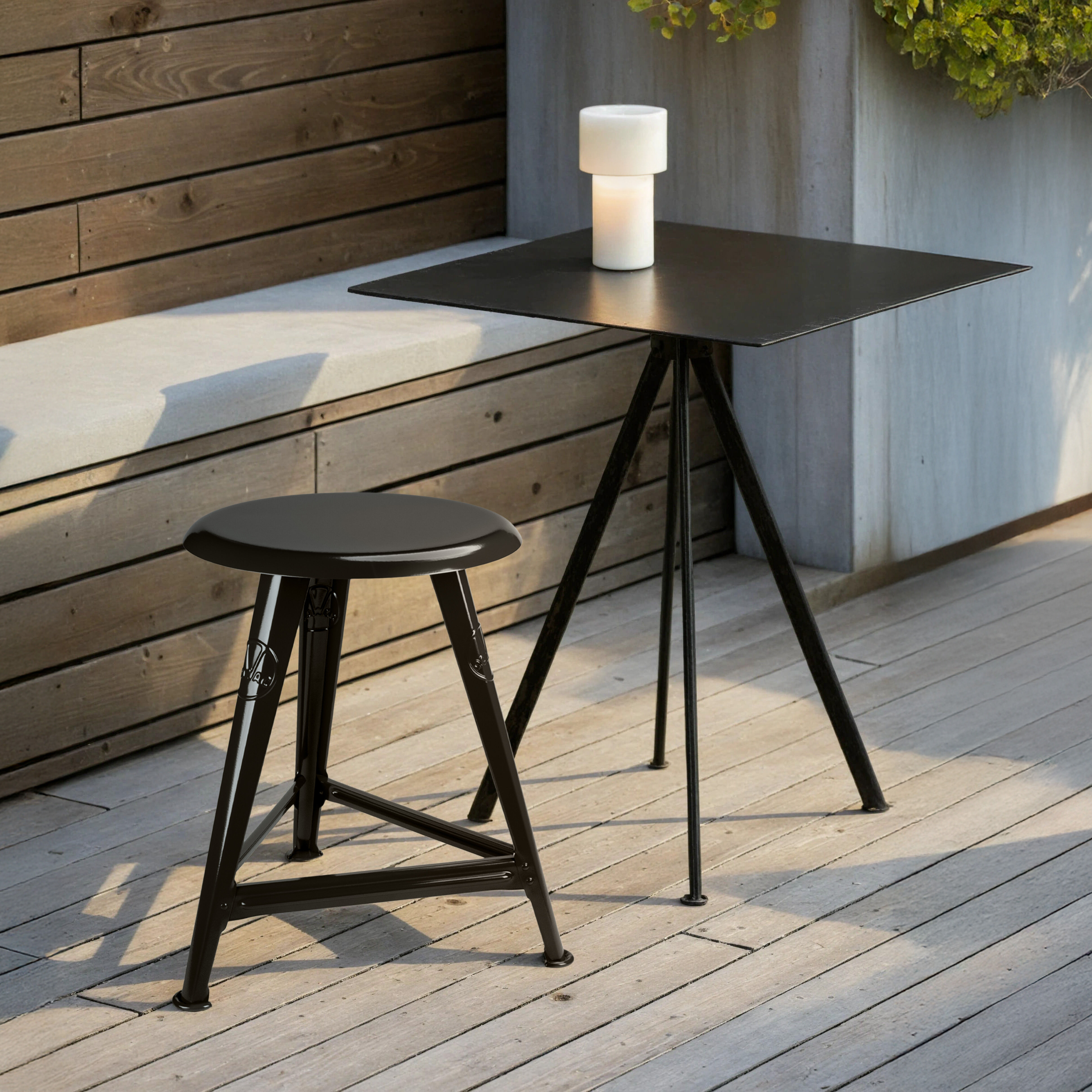 Rowac-Schemel »Just Steel« Schwarz der ROWAC GmbH steht vor einer naturbelassenen Holz-Sitzbank auf heller Holzterrasse. Der kaltgewalzte, schwarz glänzend pulverbeschichtete Stahl-Schemel präsentiert sich minimalistisch und industriell. Daneben ein moderner schwarzer Metalltisch mit weißer Dekolampe. Die Umgebung wirkt sonnig, ruhig, hochwertig und einladend.