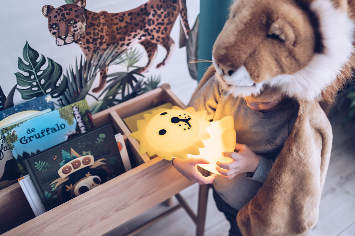 Ein Kind hält ein leuchtendes Nachtlicht in Form eines Löwen in einem Raum, der mit Kinderbüchern und einer Löwenkostüm-Dekoration geschmückt ist.