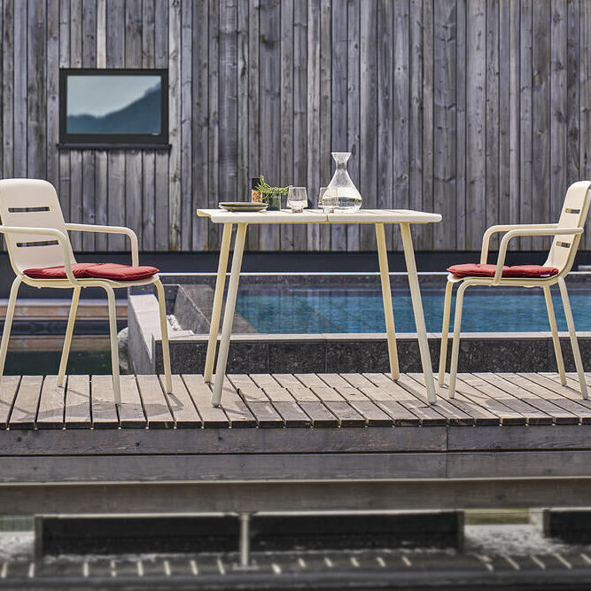 Ein Terrassentisch »VAPIO« 90x90 cm mit zwei Stühlen, die rote Sitzkissen haben, auf einem Holzdeck neben einem Pool. Im Hintergrund befindet sich ein Gebäude mit einer Holzfassade.