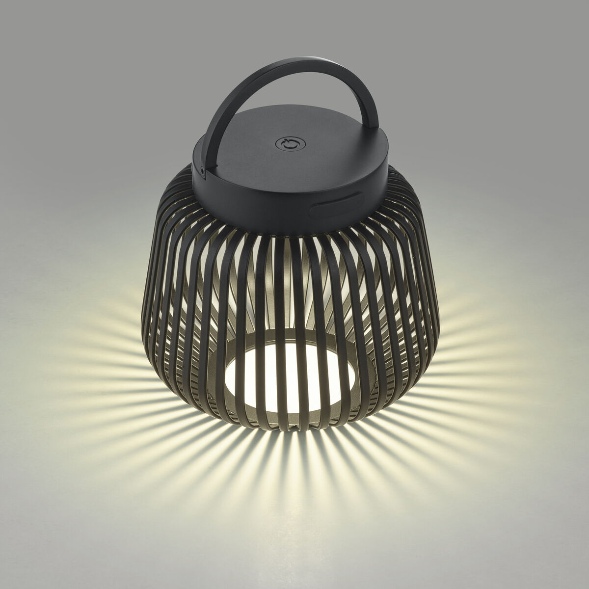 Die abgebildete Leuchte »LITO« von blomus in Granite Grey besitzt ein rundes, offenes Gitterdesign mit breiten, senkrechten Streben. Durch das LED-Licht im Inneren entstehen strahlenförmige Licht- und Schattenspiele auf dem Untergrund. Oben befindet sich ein Henkel zum Tragen.