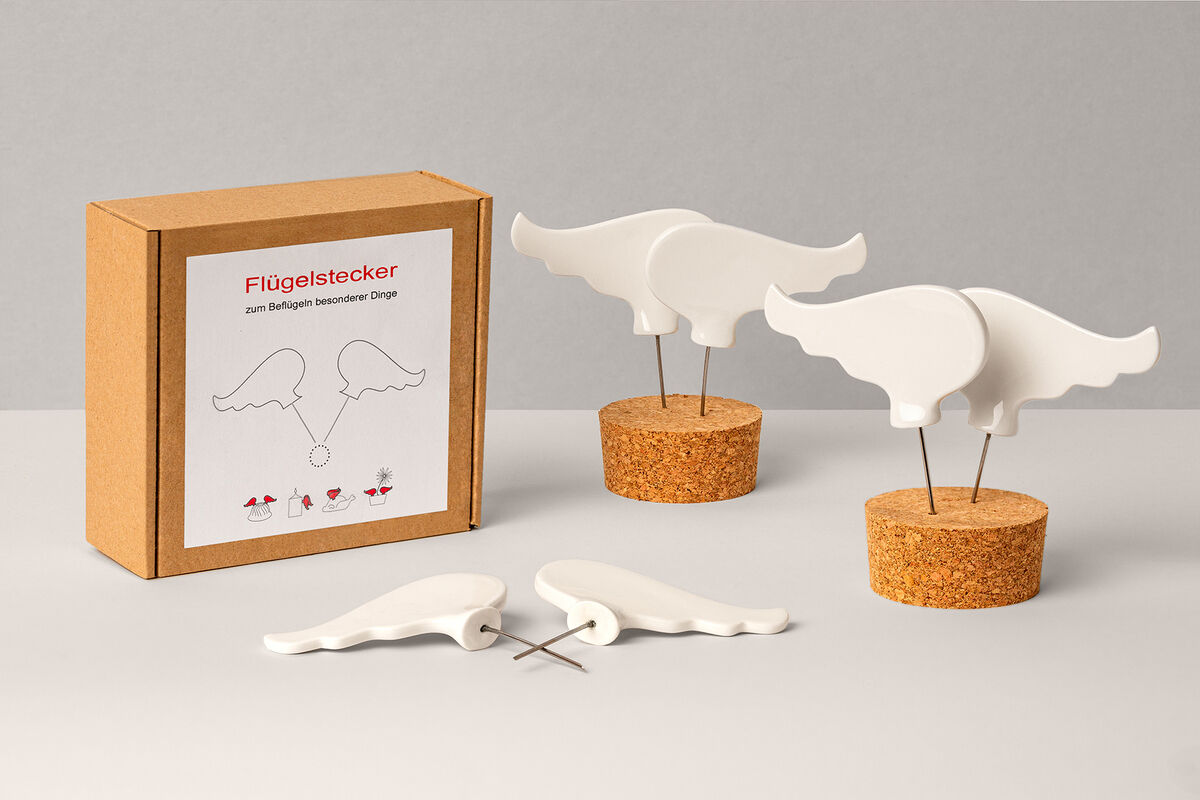 Flügelstecker Set aus Porzellan, bestehend aus zwei weißen Flügeln, die in Korkbasen gesteckt sind. Die Verpackung aus Karton ist ebenfalls zu sehen.