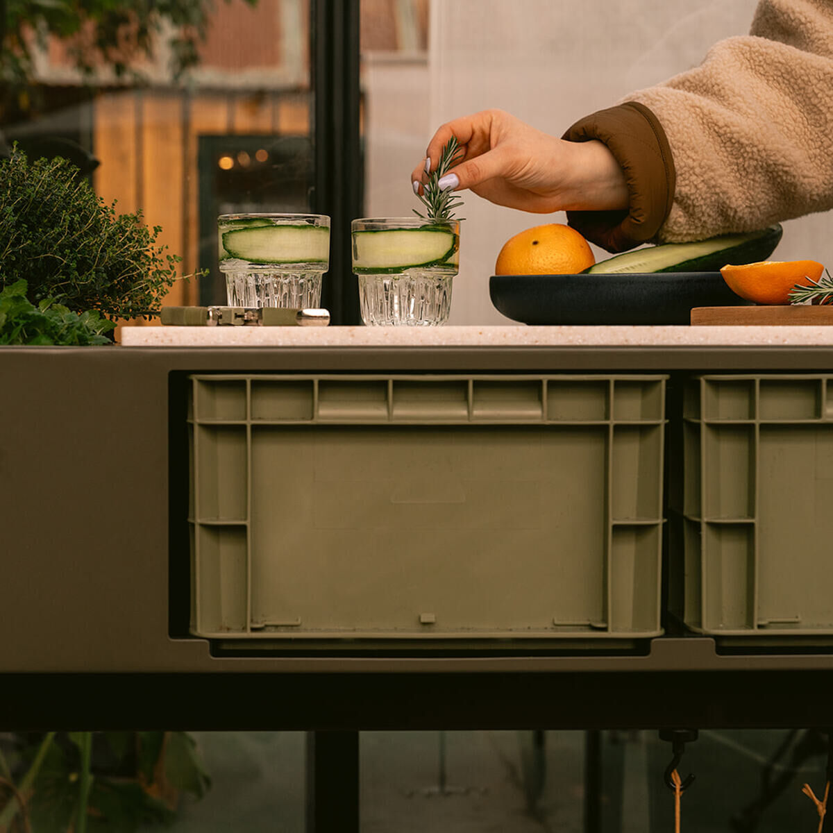 Detailreiche Aufnahme eines grünen Pflanztischs »Gardenworks Station« von Weltevree. Darauf Kräuter und gefüllte Gläser, daneben eine Schale mit Obst. Im Vordergrund zwei grüne Kisten, eine Hand dekoriert ein Getränk mit Kräutern. Draußen- oder Innenbereich.