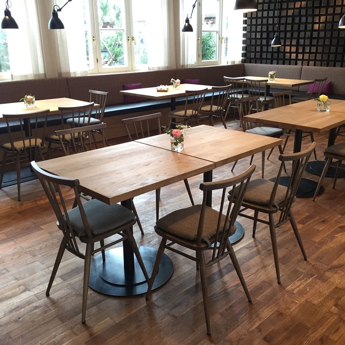 Ein stilvolles Esszimmer mit mehreren grau gebeizten Ercol »All Purpose Chair«-Stühlen, die um einfache Holztische herum angeordnet sind. Die Stühle verfügen über gepolsterte Sitze und vertikal gerahmte Rückenlehnen. Der Raum ist mit Holzboden und einer gemütlichen Sitzbank mit Kissen an der Wand ausgestattet.