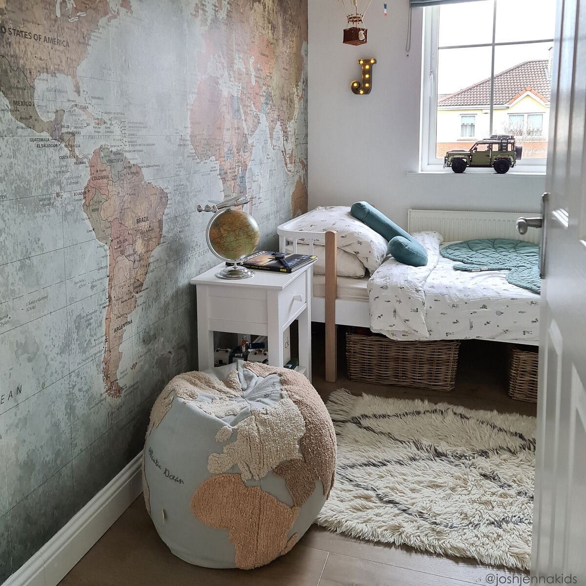 Gemütliches Kinderzimmer mit Weltkarten-Wanddekoration. Ein Sitzkissen in Form eines Weltkarten-Poufs steht neben einem kleinen weißen Tisch. Farbenfrohe Heißluftballons hängen an der Decke.