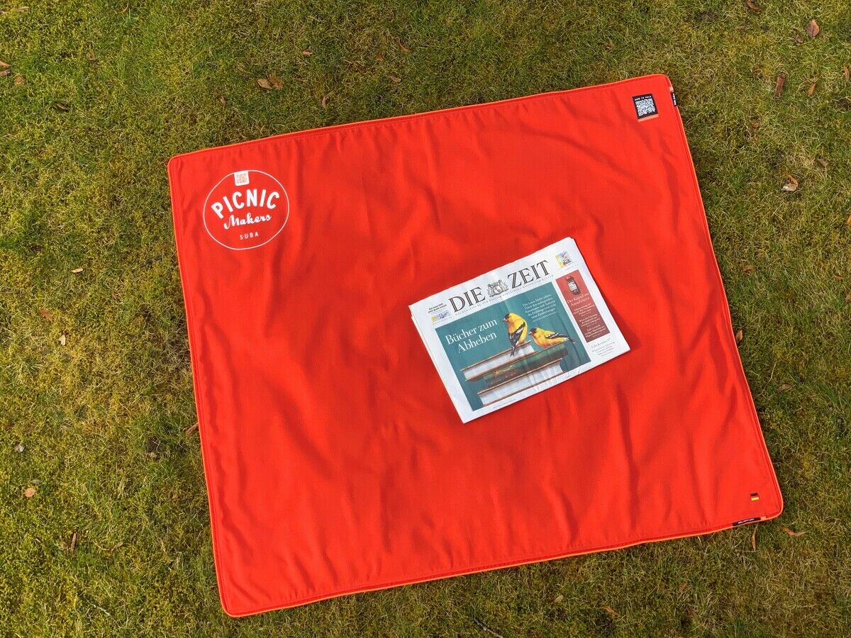 Rote Picknickdecke, quadratisch, mit der Aufschrift 'Picnic Maker', auf grünem Rasen liegend, darauf liegt eine Zeitung.