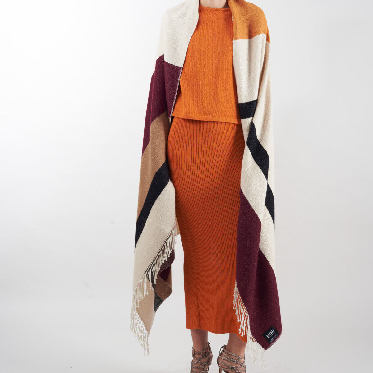 Eine Person trägt ein Cape namens "Alvar" aus Alpakawolle. Das Cape ist in den Farben Beige, Bordeauxrot, Schwarz und Weiß gestaltet. Die Person trägt darunter ein oranges Oberteil und einen passenden orangefarbenen Rock.