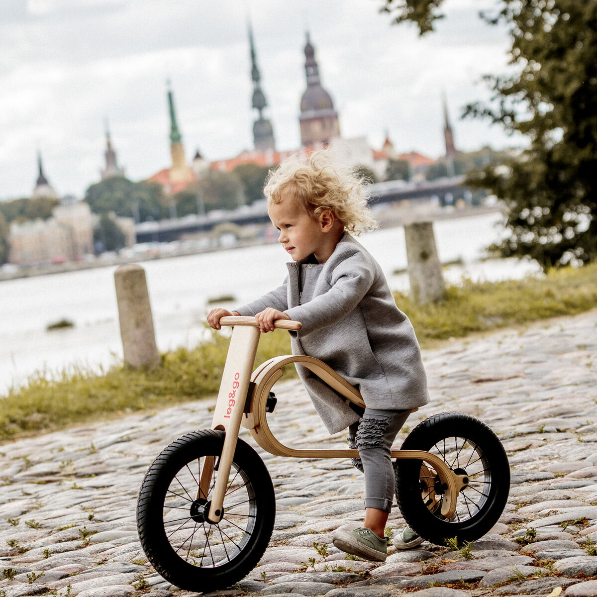 Ein Kleinkind fährt auf einem Balance Bike 3in1 aus Holz auf einem gepflasterten Weg entlang. Im Hintergrund sind eine Flusslandschaft und die Silhouette einer Stadt mit mehreren Kirchtürmen sichtbar. Das Laufrad hat schwarze Reifen und einen natürlichen Holzrahmen. Die Szene spielt im Freien, bei Tageslicht.