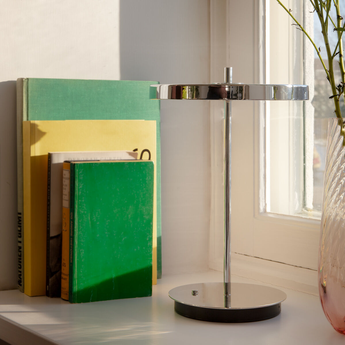 Eine moderne Tischlampe Asteria Move aus poliertem Stahl steht auf einer weißen Fensterbank neben mehreren farbigen Büchern und einer Glasvase mit gelben Blumen. Sonnenlicht fällt durch das Fenster und unterstreicht das elegante, minimalistische Design der Leuchte.