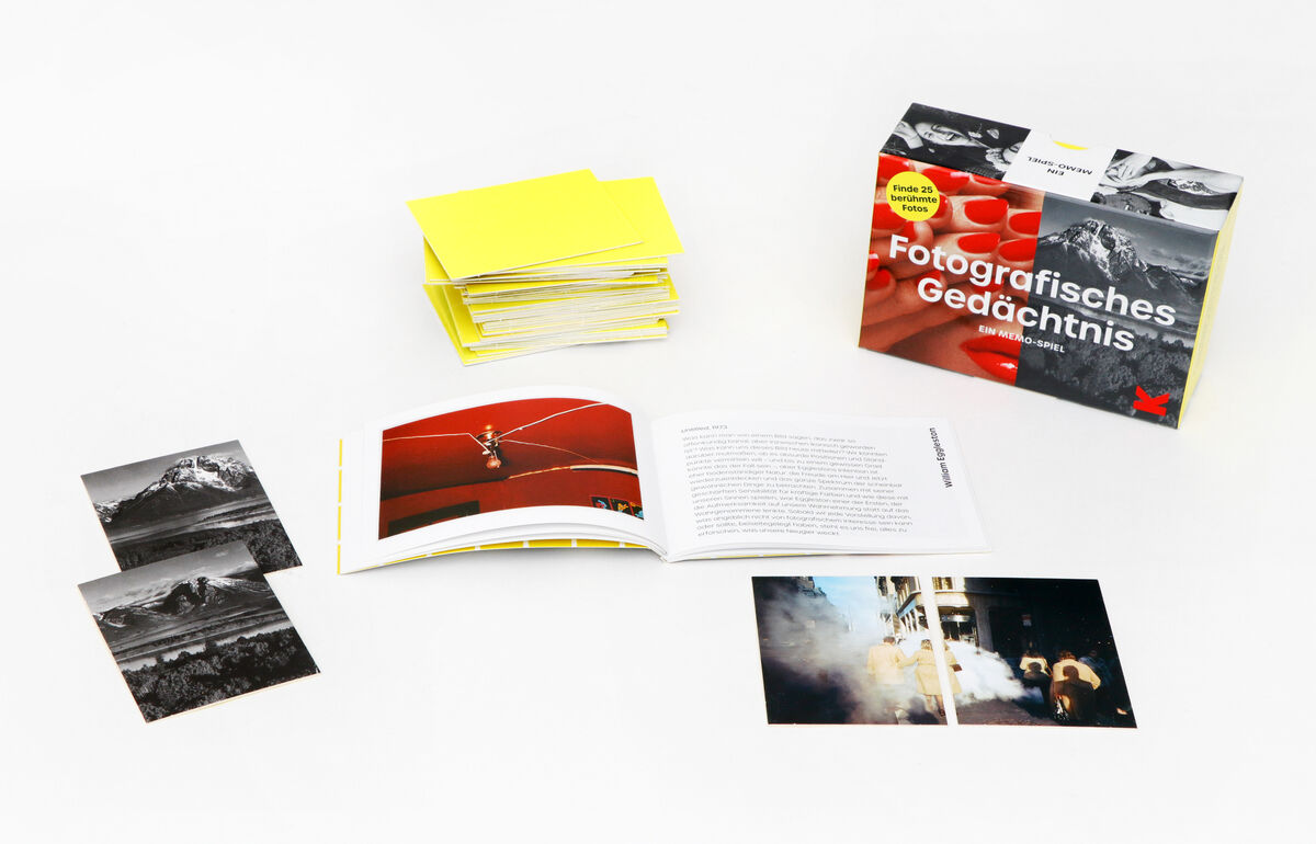 Ein Memo-Spiel mit dem Titel »Fotografisches Gedächtnis«. Auf dem Bild sind verschiedene Bestandteile des Spiels zu sehen: ein Spielkartenstapel in Gelb, ein geöffnetes Buch mit Fotografien und Text sowie mehrere lose Fotokarten in Schwarz-Weiß und Farbe. Im Hintergrund ist die Verpackung des Spiels abgebildet.
