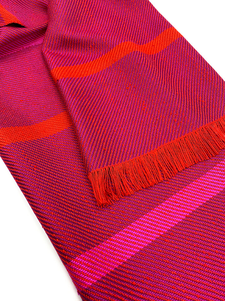 Nahaufnahme eines gewebten Seidenschals in Fuchsia und Rot mit Fransen an einem Ende. Der Schal zeigt ein dezentes Muster in verschiedenen Rottönen.