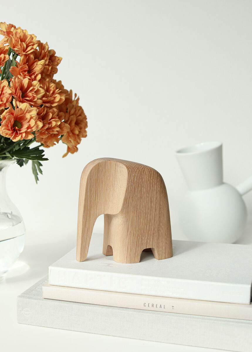 Eine minimalistische Holzfigur in Form eines Elefanten aus Eiche steht auf einem Stapel von weißen Büchern. Daneben steht eine Vase mit orangefarbenen Blumen und eine weiße Kanne. Der Hintergrund ist hell und schlicht.