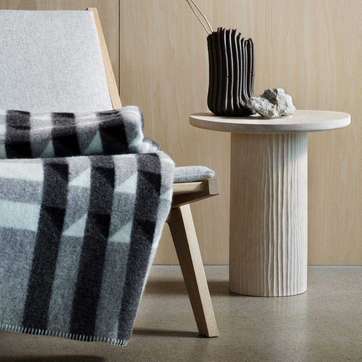 Eine gestreifte Lammwolldecke mit geometrischem Muster liegt auf einem modernen Holzstuhl. Ein kleiner Rundtisch neben dem Stuhl trägt eine dunkle Vase mit dekorativen Zweigen. Hintergrund ist eine Holzpaneelwand.