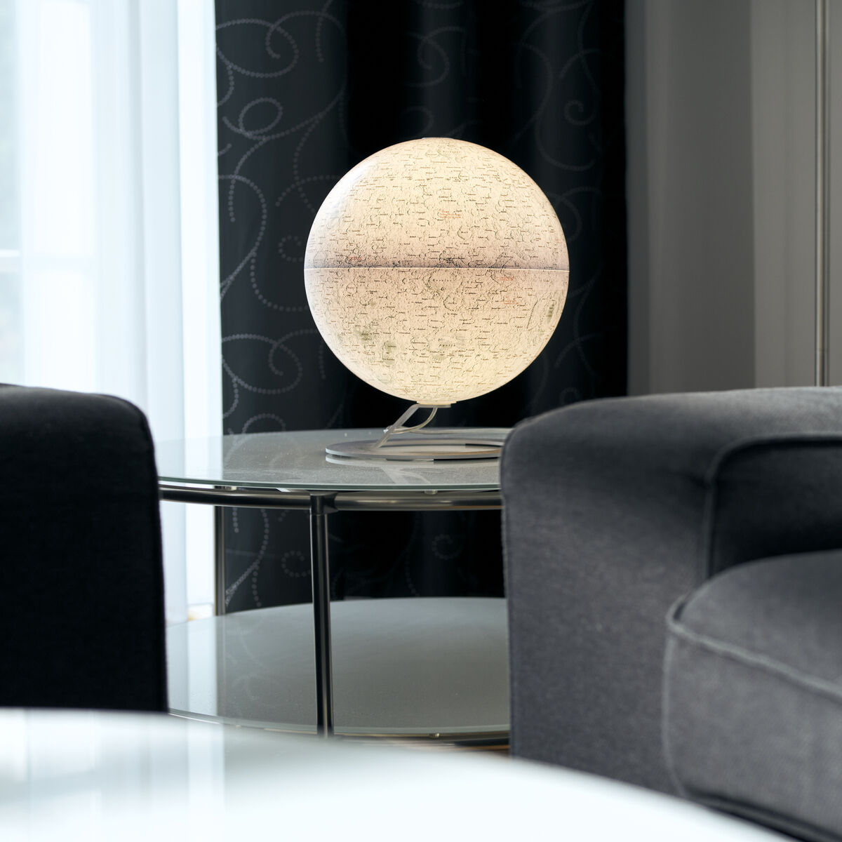 Ein Leuchtglobus von National Geographic mit dem Thema Mond, platziert auf einem Tisch in einem modernen Wohnzimmer. Der Globus leuchtet angenehm und erzeugt eine stimmungsvolle Beleuchtung. Die Umgebung ist in Schwarz- und Grautönen gehalten.