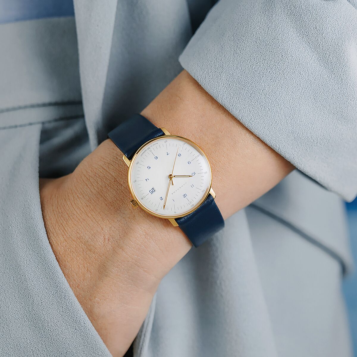 Zu sehen ist eine elegante Damenuhr der Marke Junghans, Modell ZEIT Sonderedition »max bill Damen« mit weißem Zifferblatt, goldfarbenem Edelstahlgehäuse und dunkelblauem Lederarmband am Handgelenk einer Person. Stilvoller, schlichter Look.