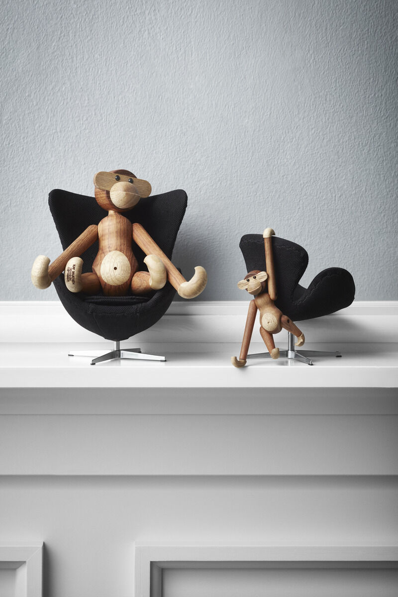 Zwei Holzfiguren in Affenform, entworfen von Kay Bojesen. Die Figuren sind auf einem Regal platziert, eine sitzt in einem schwarzen Stuhl und die andere steht, ebenfalls in einem schwarzen Stuhl. Der Hintergrund ist eine helle Wand.