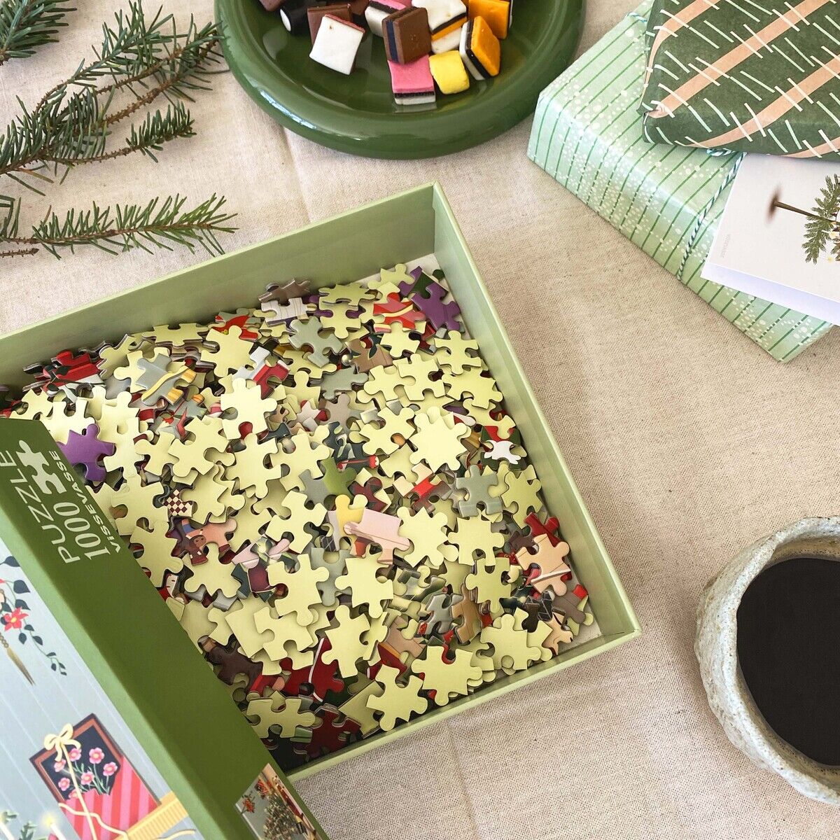 Geöffnetes Puzzle von VISSEVASSE mit 1000 Teilen im Karton, daneben ein grün gemusterter Geschenkkarton, ein runder grüner Teller mit buntem Konfekt sowie Tannenzweige auf beigem Tuch. Die Szene wirkt einladend und festlich dekoriert.