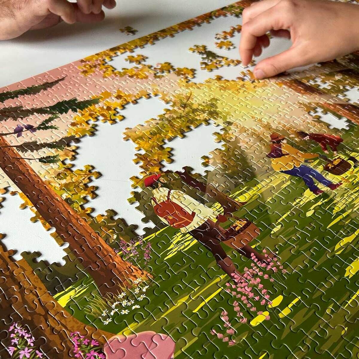 Mehrere Hände setzen ein 1000-Teile-Puzzle mit einer bunten Waldszene zusammen, in der Menschen durch einen Wald voller gelber Blätter und blühender Blumen spazieren.