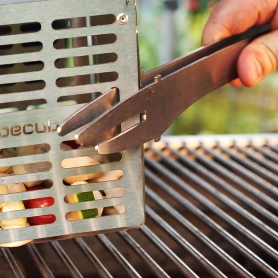 Eine Person hält eine Grillzange und positioniert damit einen Grillkorb namens 'The Cube' über einem Grillrost. Der Grillkorb enthält buntes Gemüse. Die Szenerie ist auf ein Außengrill-Setup fokussiert.