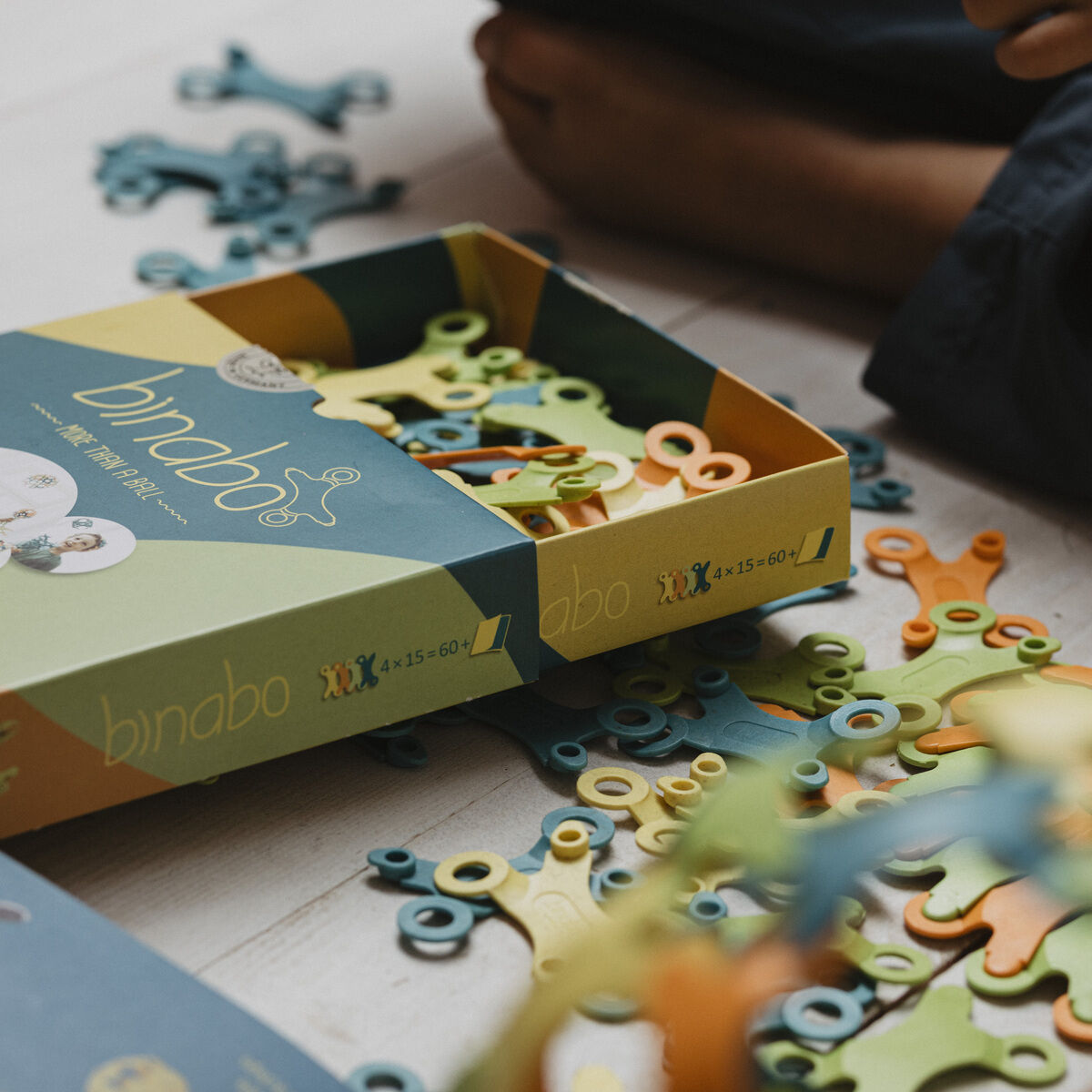 Spiel »Binabo« 60 Teile - Bunte, recycelbare Kunststoffteile in einer offenen Schachtel, umgeben von Kinderhänden, die darauf hinweisen, dass sie bald zum Spielen verwendet werden.
