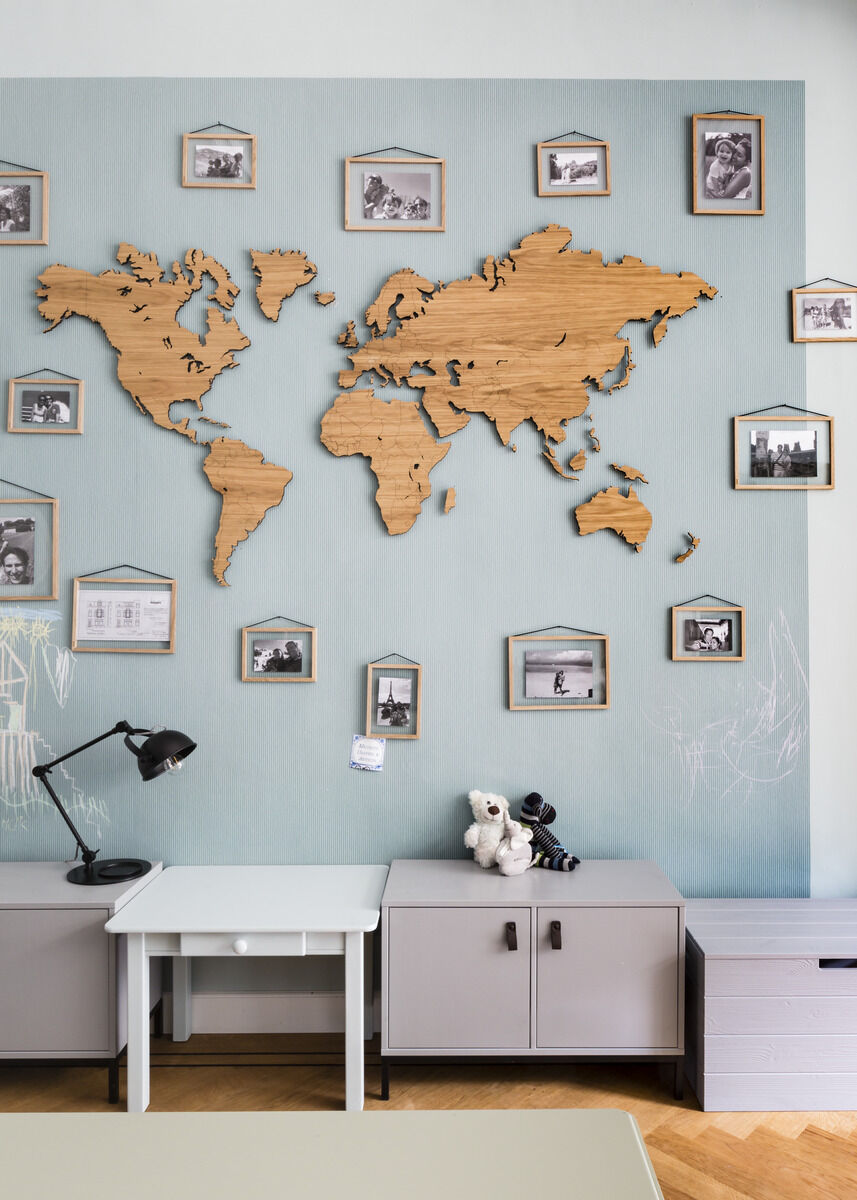 Weltkarte aus Eichenholz an einer hellgrauen Wand, umgeben von gerahmten Fotos. Darunter befinden sich ein Schreibtisch mit einer schwarzen Lampe, einige Spielzeuge und Schränke.