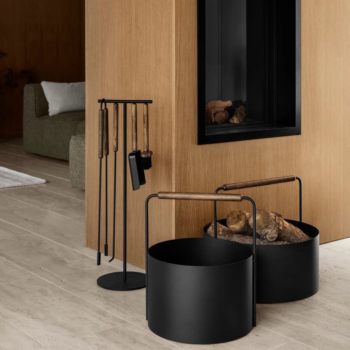 Modernes Kaminbesteck-Set aus schwarzem Metall und Holzgriffen steht neben einem Kamin mit Holzlager, vor einer hellbraunen Holzverkleidung und hellen Dielen in einem stilvoll eingerichteten Wohnzimmer.
