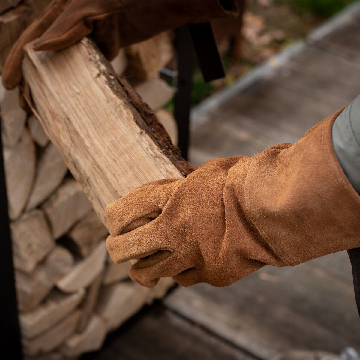 Robuste, braune Arbeitshandschuhe aus Rindleder greifen ein Stück Holz; Hintergrund zeigt gestapeltes Holz.