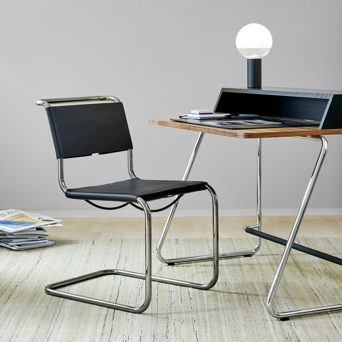 Ein moderner Schreibtisch »S1200« von Thonet aus Holz und Metall. Der Schreibtisch hat ein elegantes, minimalistisches Design mit klaren Linien. Auf der Tischplatte steht eine Lampe. Neben dem Schreibtisch steht ein Stuhl mit schwarzer Sitzfläche und metallenen Beinen. Auf dem Boden liegt ein heller Teppich, und im Hintergrund sind Zeitschriften gestapelt.