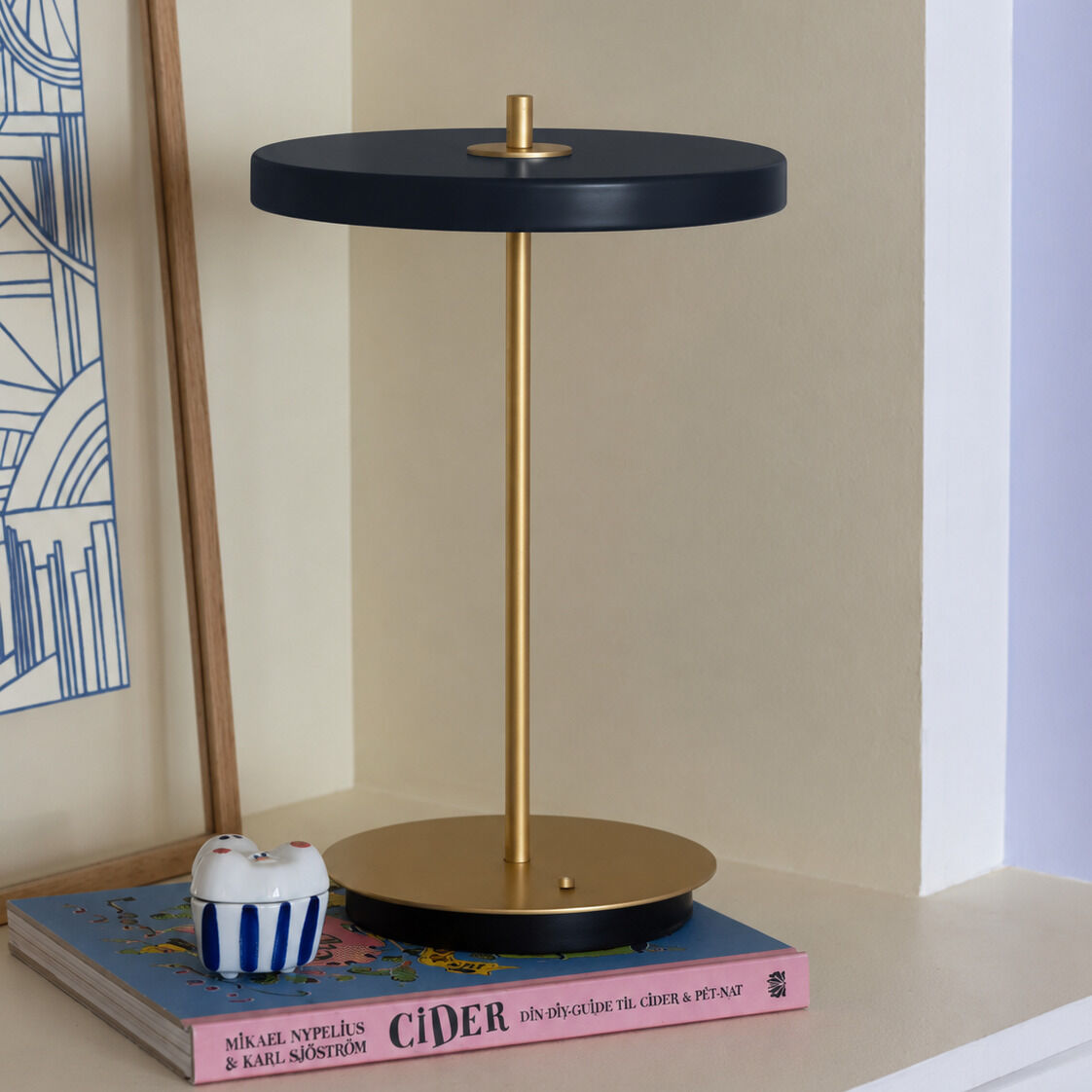 Die kabellose Tischleuchte »Asteria Move« von Umage in Midnight Blue und Messing steht auf einem Regal neben einer rosa Buchbasis. Der minimalistische Leuchtenkörper aus Metall setzt edle Akzente, die runde Form harmoniert mit modernem skandinavischem Design.