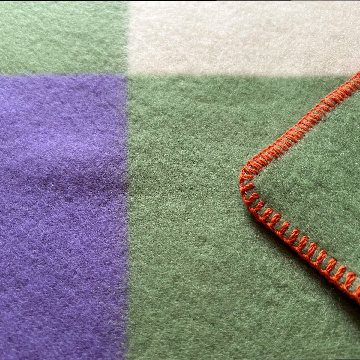 Nahaufnahme einer Decke namens 'Tricolor' mit drei verschiedenen Farben: grün, lila und beige, sowie einer orangefarbenen Naht am Rand. Die Textur wirkt weich und kuschelig.