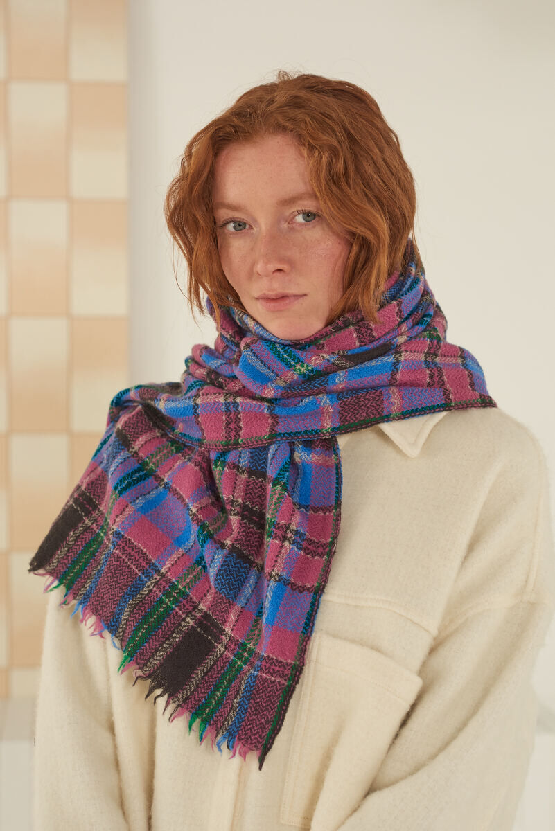 Eine Frau mit roten Haaren trägt einen Schal »No. 700« in den Farben blau, violett, grün und rosa mit Plaid-Muster. Sie hat eine cremefarbene Jacke an und steht vor einem neutralen Hintergrund.