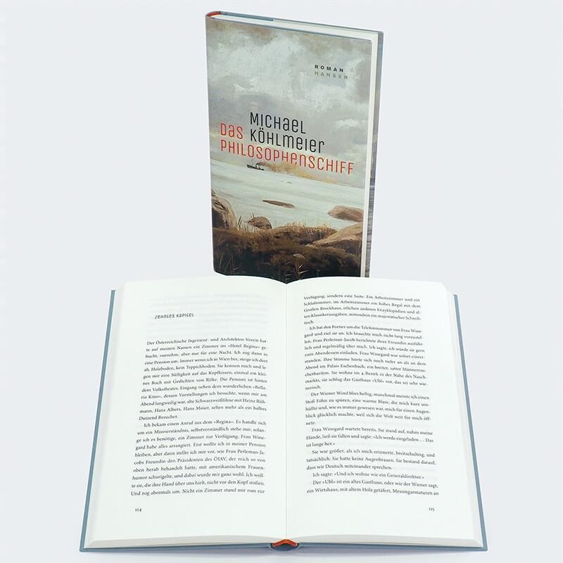 Das Bild zeigt ein Buch von Michael Köhlmeier mit dem Titel 'Das Philosophenschiff', das aufrecht steht. Davor liegt ein geöffnetes Buch mit Fließtext auf weißem Hintergrund.