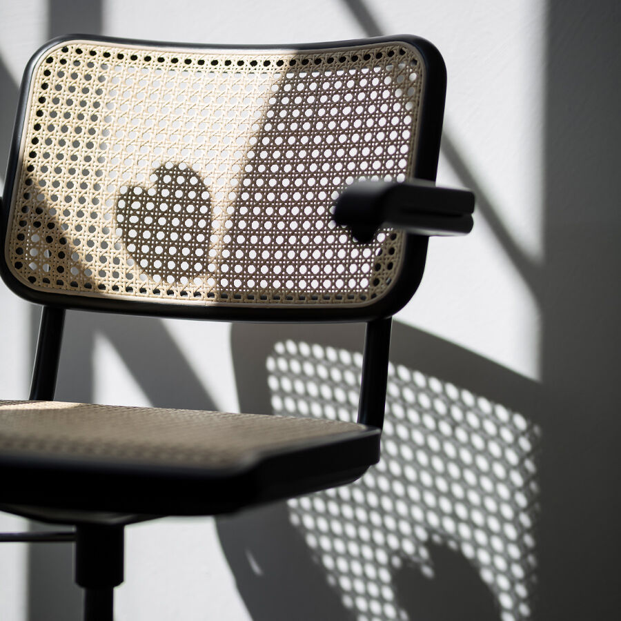 Detailaufnahme des Schreibtischstuhls »S 64 Atelier« von Thonet: Sitz und Rückenlehne aus hell gebeiztem Buchenholz mit Rohrgeflecht, das Licht wirft ein kunstvolles Schattenspiel und hebt die klaren Linien sowie das ikonische Bauhaus-Design hervor. Armlehne und Stuhlgestell sind aus glänzendem Chromstahl.