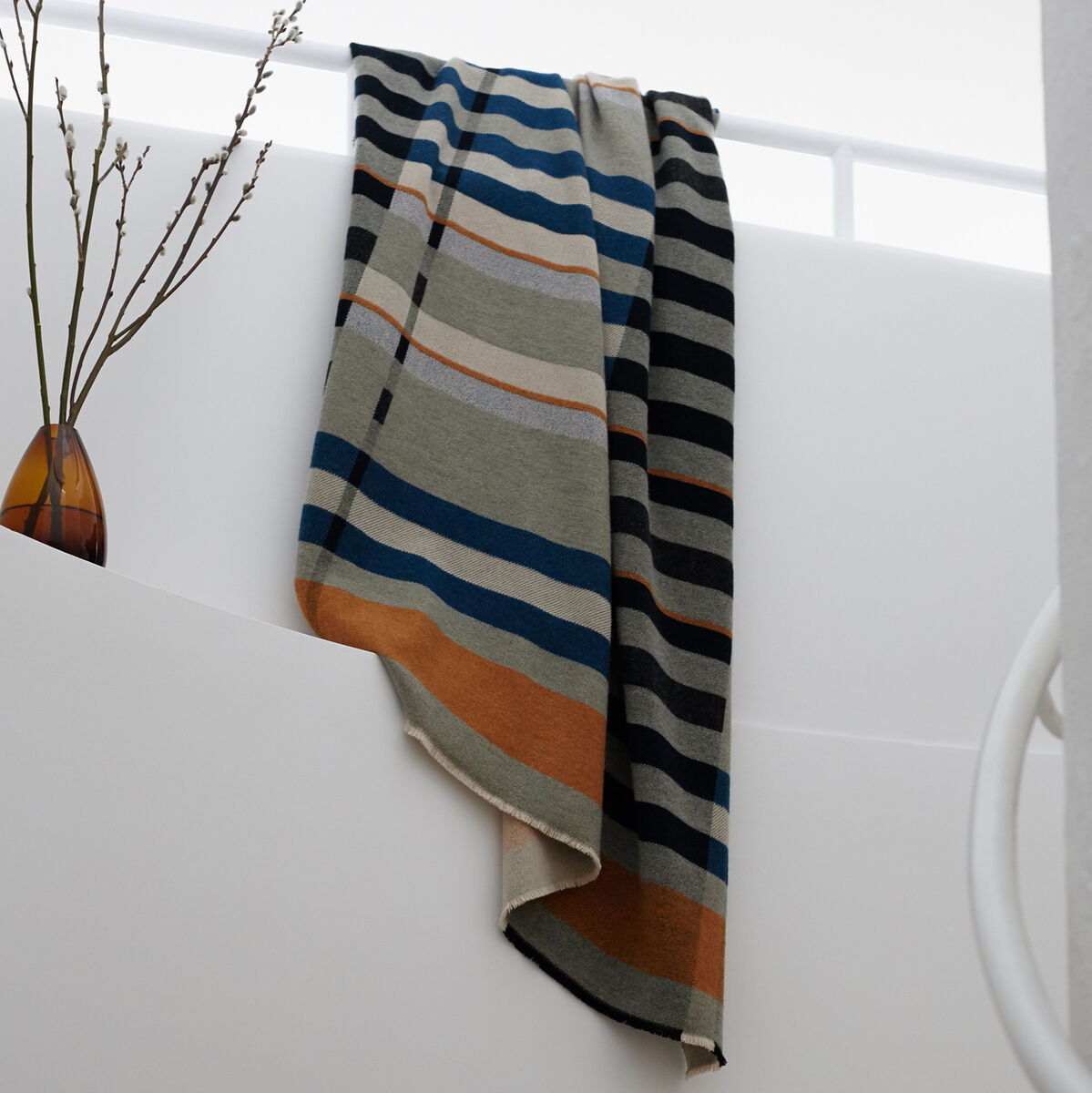 Eine gestreifte Decke mit Beige-, Blau-, Orange-, Schwarz- und Weißtönen hängt elegant von einem Geländer auf einer Treppe herab. Eine Vase mit Zweigen befindet sich auf dem Treppenabsatz in einem modernen, hellen Raum.