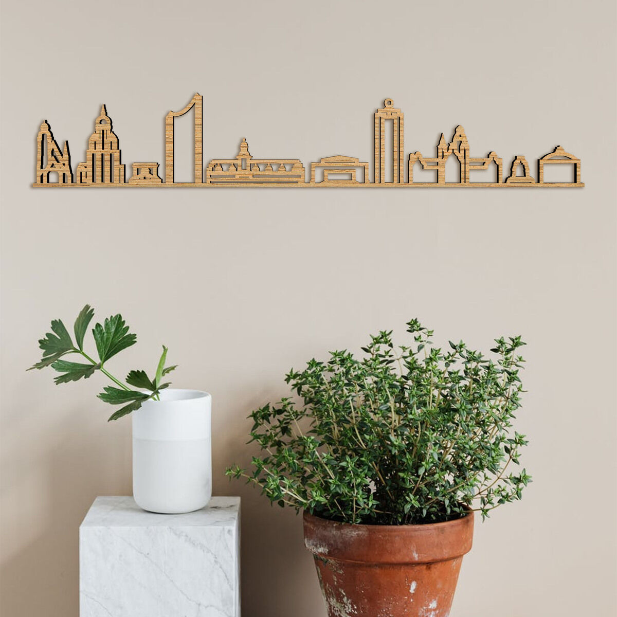 Holzschnitt der Skyline von Leipzig an einer beigen Wand. Dekoriert mit Pflanzen in einem Blumentopf und einer weißen Vase auf einem weißen Sockel.